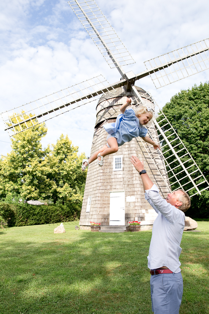 dad tosses little blonde girl in blue dress up in the air at the Beebe Windmill in Bridgehampton
