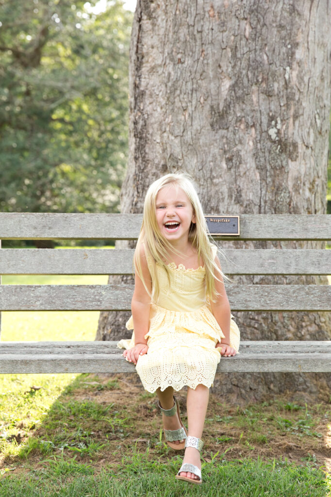 blonde girl in yellow dress sits on bench laughing at the camera