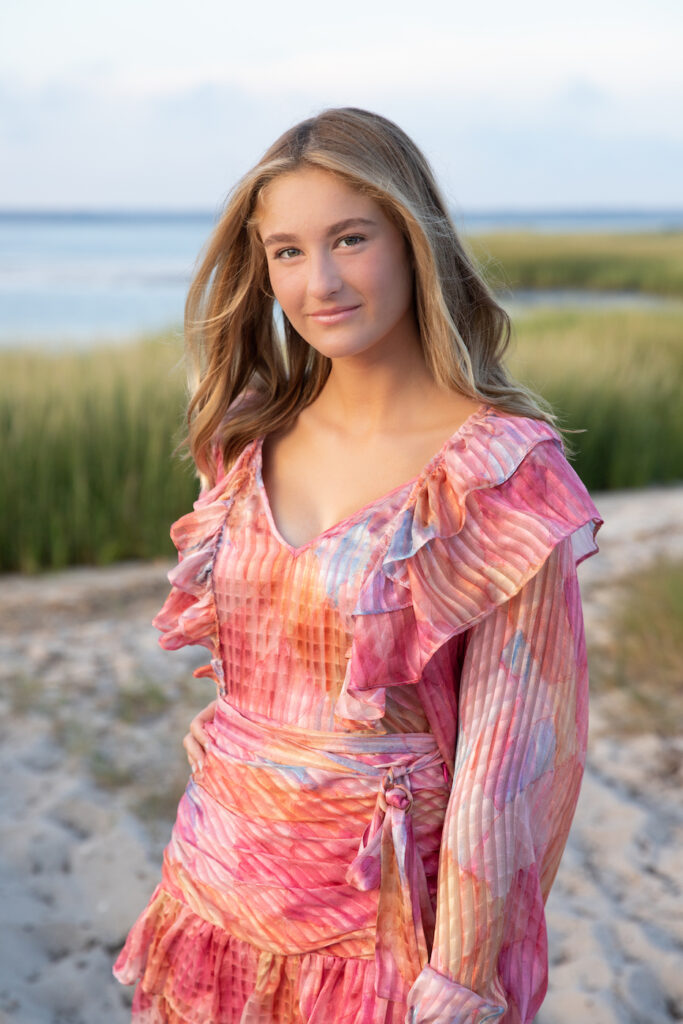 High school senior girl in pink dress looks right at the camera with beach grasses and bey behind her in Southampton