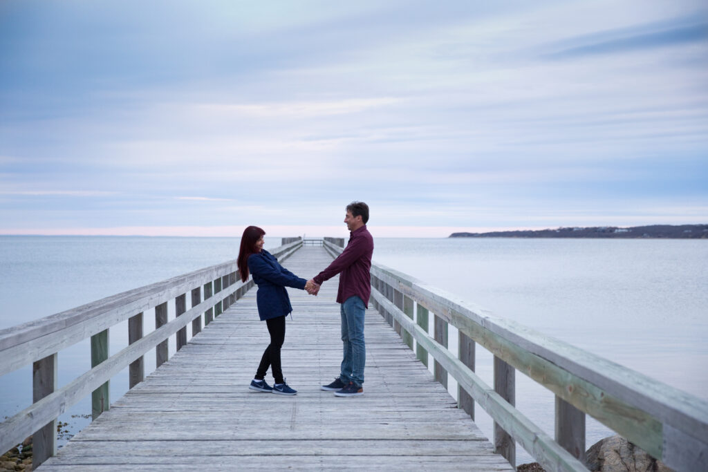 Couple celebrates their anniversary holding hands looking at each other on the dock at Navy Beach in Mountauk with the purple blue post-sunset sky in the background