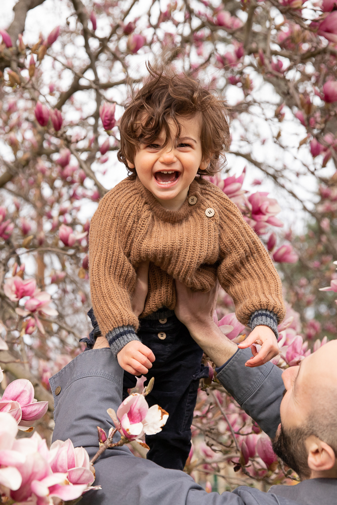 father lifts baby boy into air with magnolia tree in the background and baby boy looks right at camera with big smile