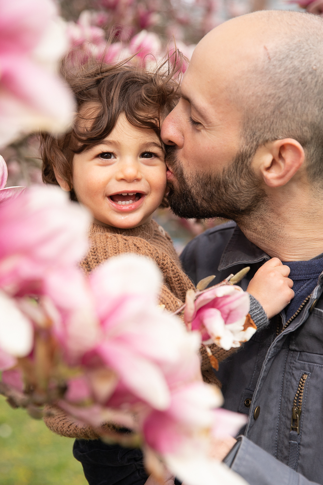 father kisses baby boy with magnolia flowers blurred in the foreground and background