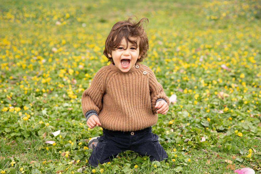 Baby boy kneels in field of yellow spring flowers with a big open mouthed smile looking right at the camera in the Hudson Valley, NY