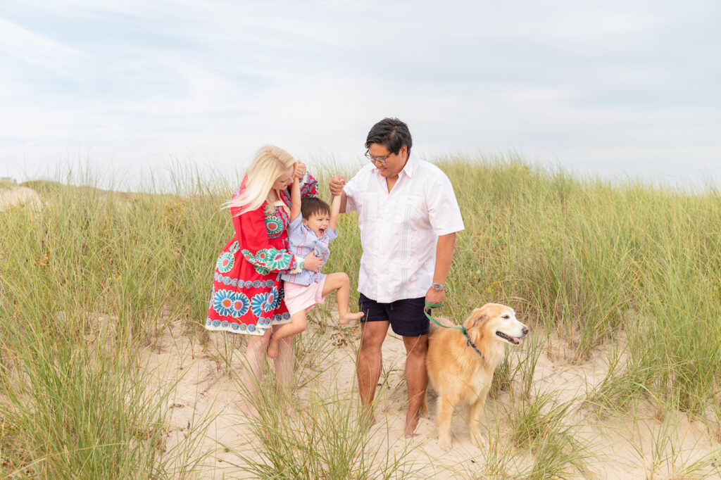 Parents lift up toddler son tickling him in the grassy dunes at Egypt Beach in East Hampton while golden retriever looks ourt to the beach