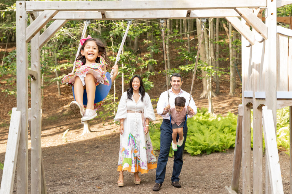 Family of four play in their East Hampton backyard on the swingset with little girl swinging towards the camera with a huge smile