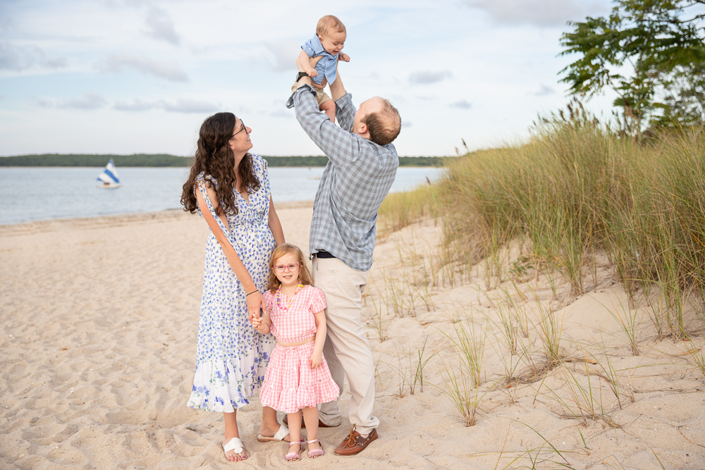 Family of 4 on Haven's Beach in Sag Harbor with a sailboat passing in the background. Dad lifts baby into the air, little girl stands looking at the camera and mom holds little girl's hand while looking up at baby.,