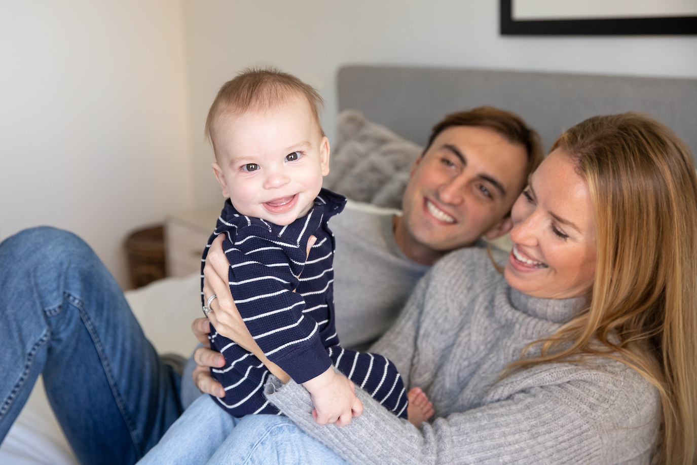 baby boy sits on mom's knee on the bed smiling towards camera at NYC family photo shoot