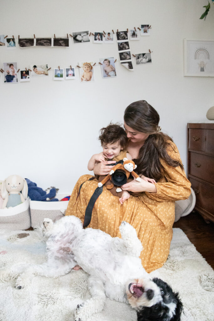 Westchester photographer Laura Volpacchio Simon sits at home with her baby on her lap, holding camera and laughing at dog rolling over on the floor.