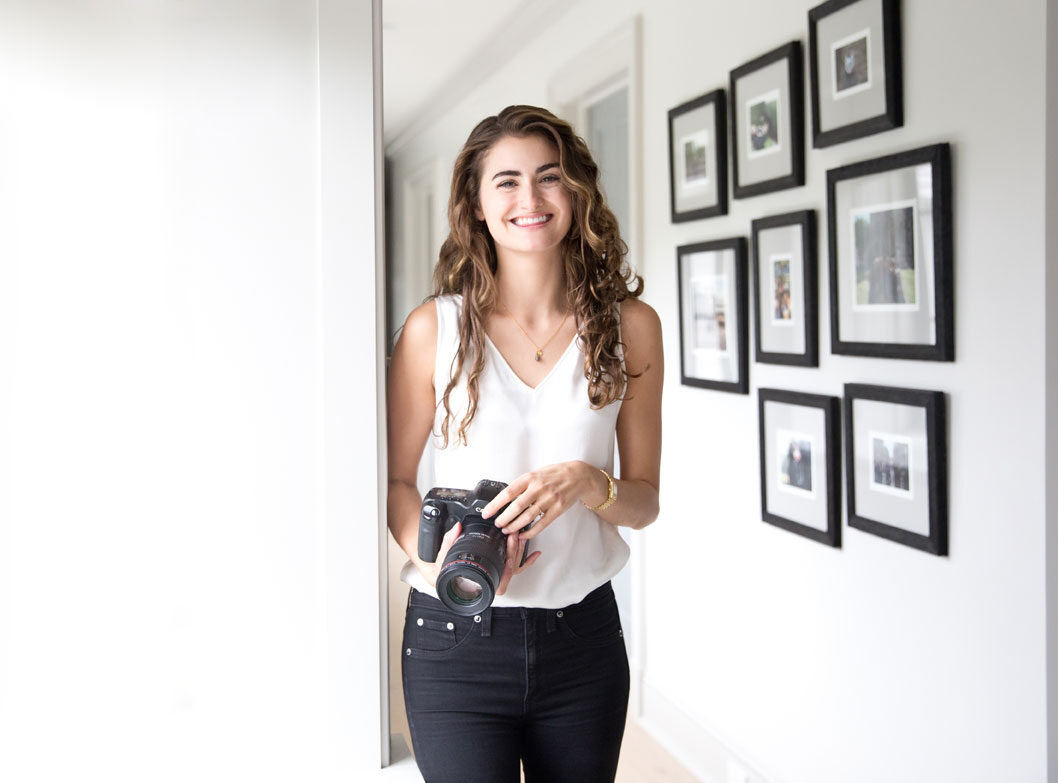 New York photographer Laura Volpacchio Simon holds camera smiling with a wall of framed photographs behind her.