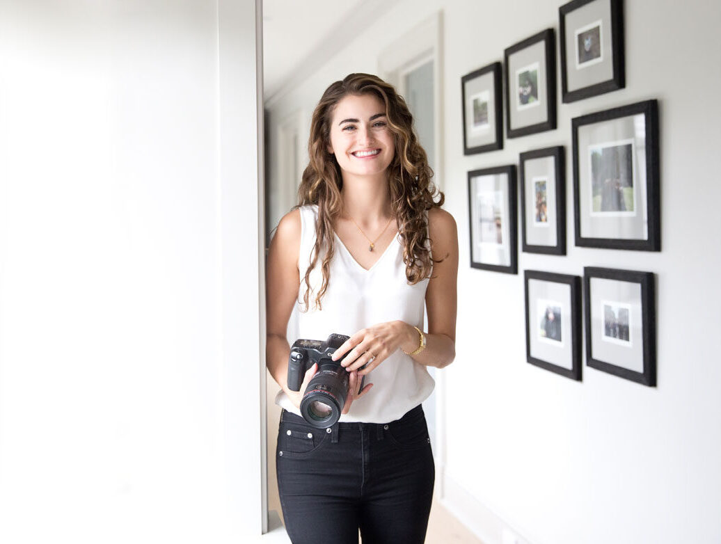 New York photographer Laura Volpacchio Simon holds camera smiling with a wall of framed photographs behind her.